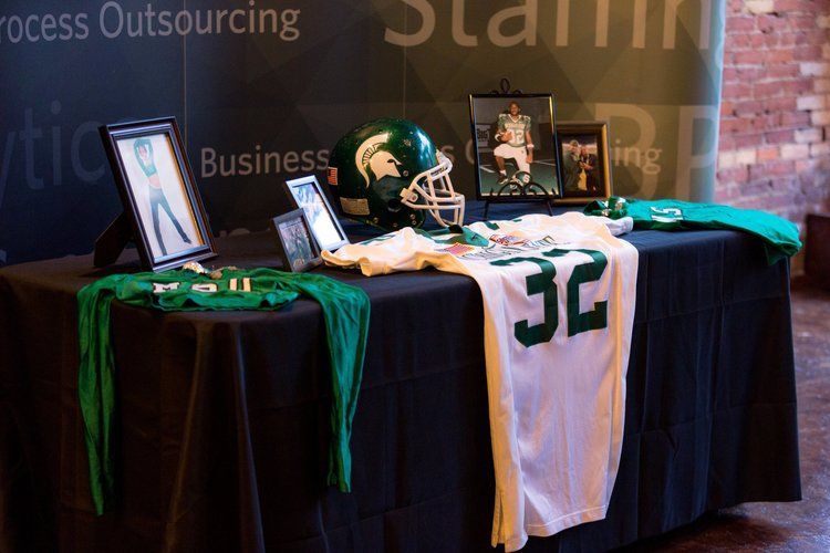 Table with sports memorabilia: green football helmet, jersey, photos. Black tablecloth, brick wall background.