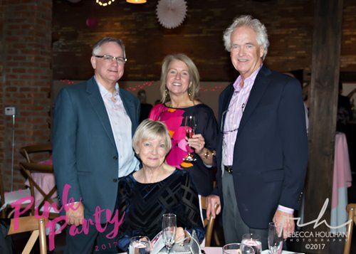 Four people at a pink-themed event, posing for a photo.  A woman sits while others stand; smiling.