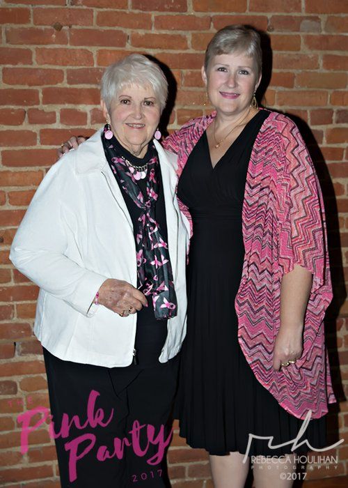 Two women pose together, smiling, near a brick wall. One wears a white jacket and a pink scarf, the other a black dress and pink shawl.