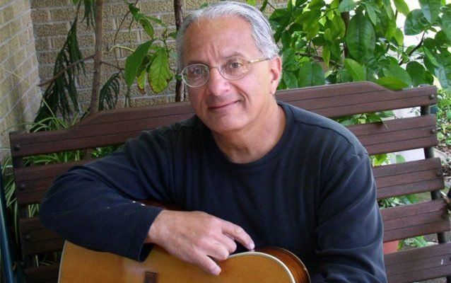 Man with glasses and guitar sits on a wooden bench, leaning against it.