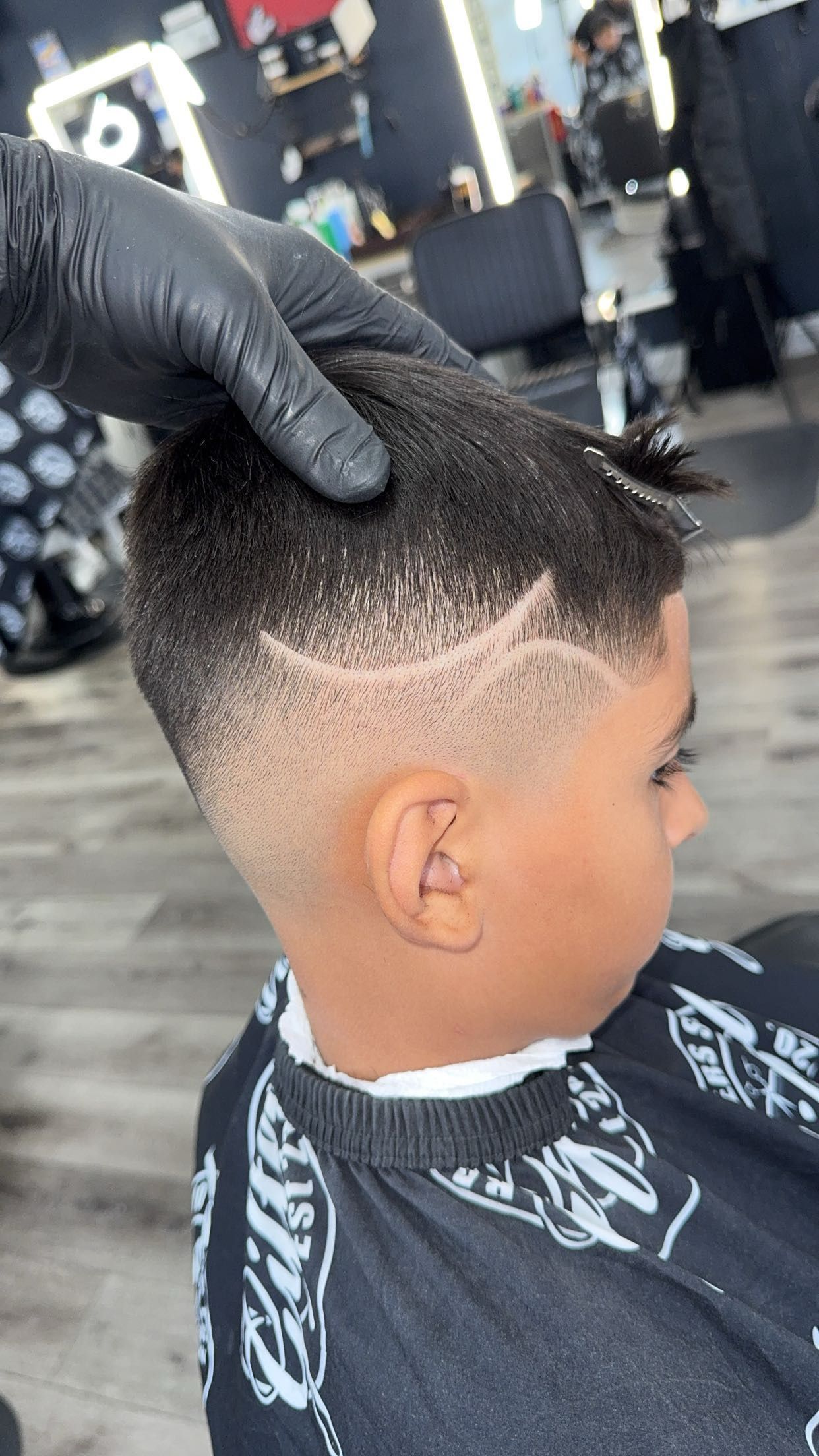 Boy with a faded haircut and etched design. Black-gloved hand is touching his hair in a barber shop.