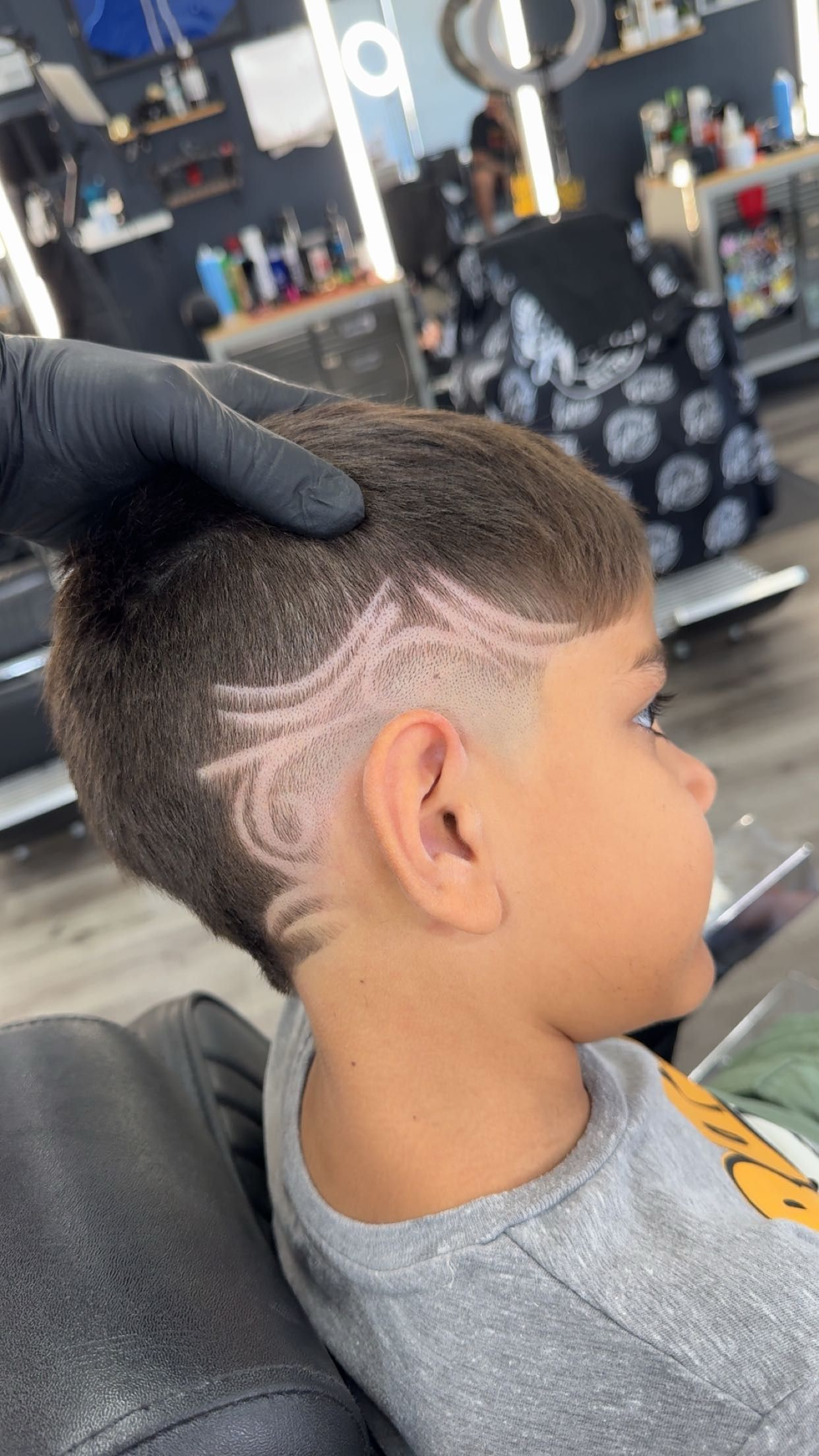 Young boy with a patterned hair design at a barbershop, held by a gloved hand.