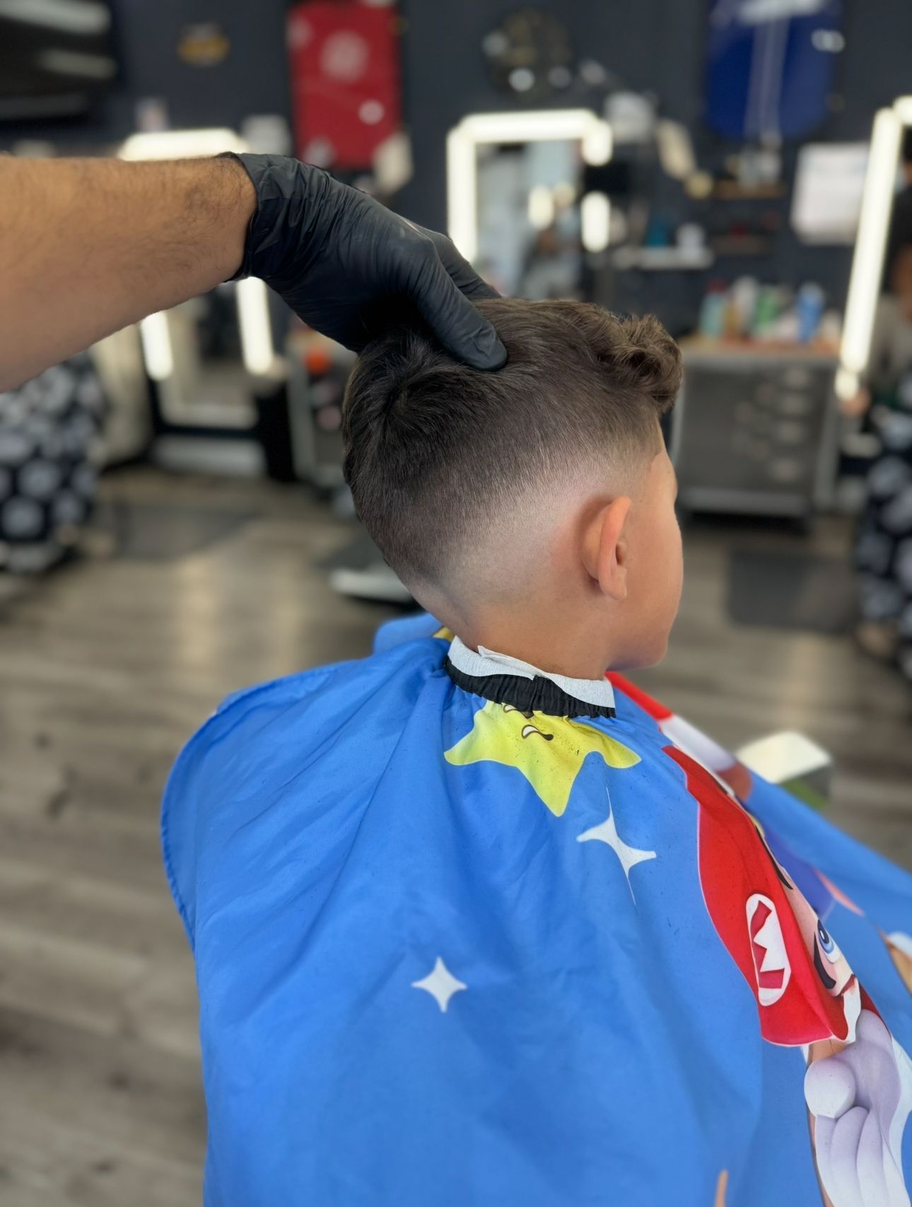 A barber gives a boy a fade haircut. Boy is wearing a blue cape with Mario characters.