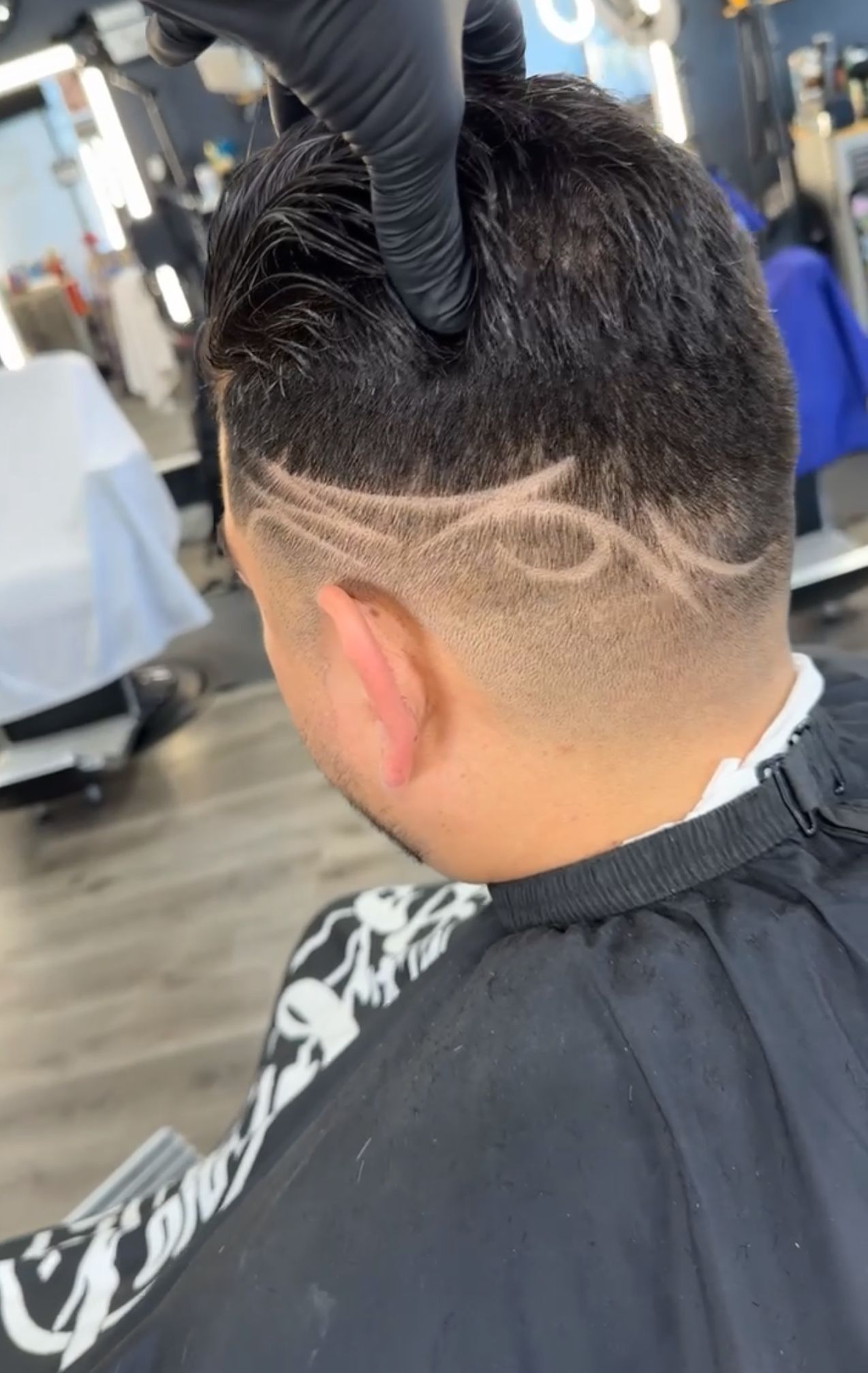 Man's hair with intricate design shaved on the side. Barber's hands in black gloves touch the head, indoors.
