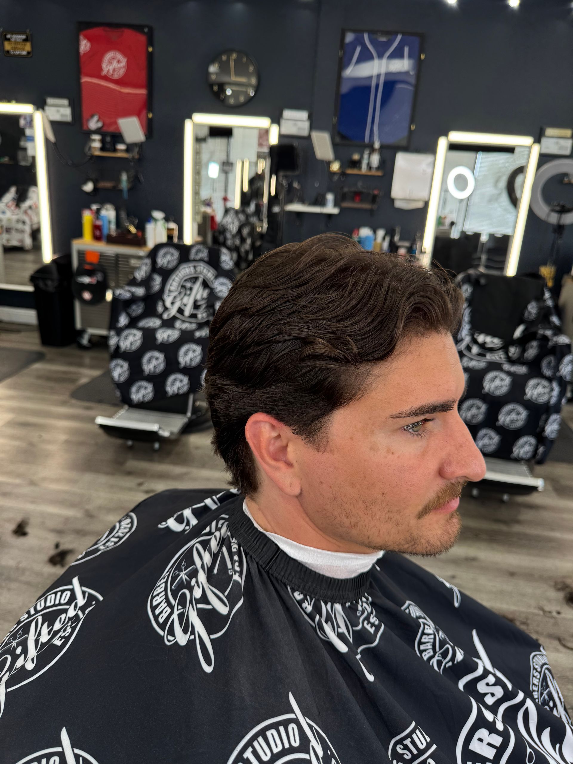 Man in barber's chair with new haircut, dark hair, and mustache. In a barbershop with mirrors and chairs.