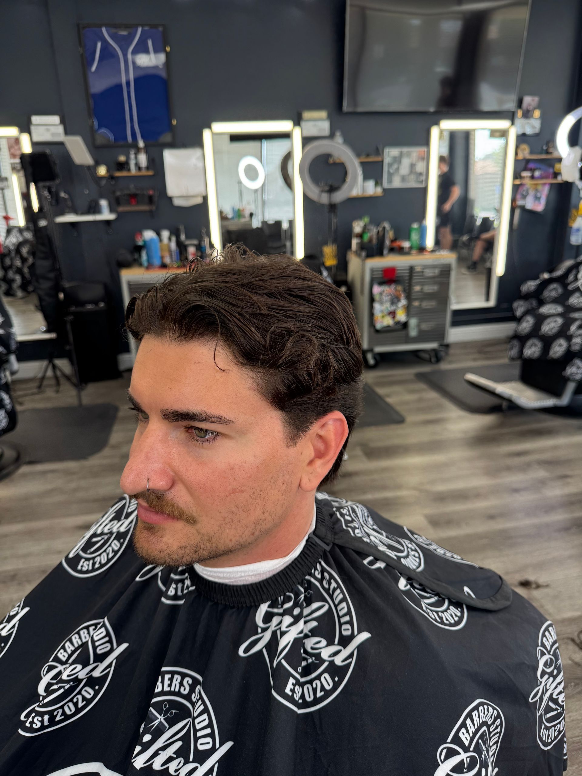 Man in barber's chair; dark hair, mustache, gray barber cape. Barbershop interior.