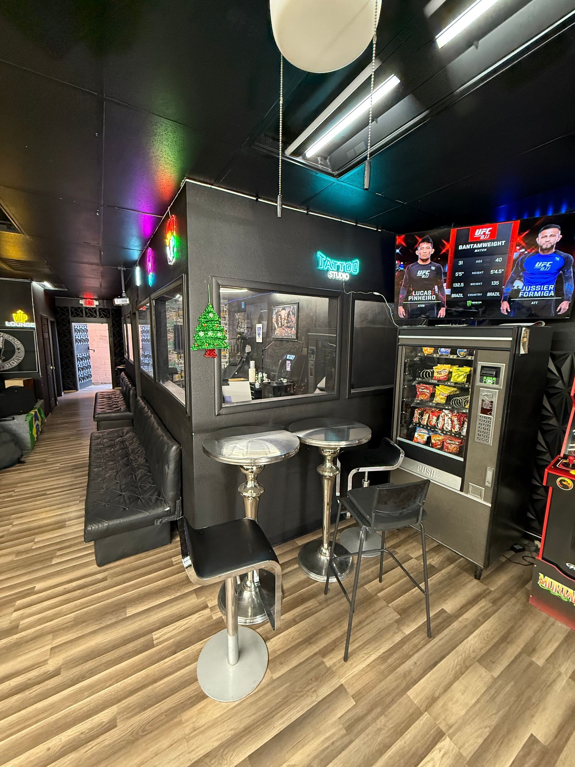Interior of a black-themed game room with neon lights, arcade machines, seating, and a vending machine.