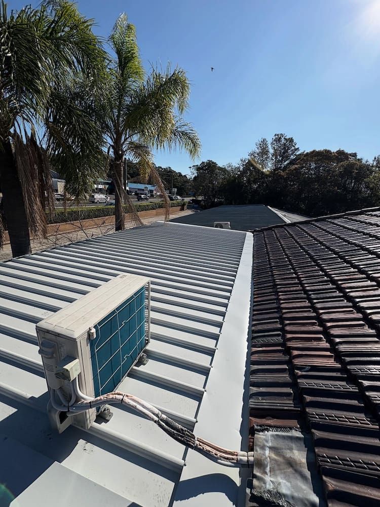 An outdoor air conditioning unit sits on a light gray metal roof next to a dark tiled roof under a clear blue sky — Crystal Clear Plumbing & Roofing in Wollongong, NSW