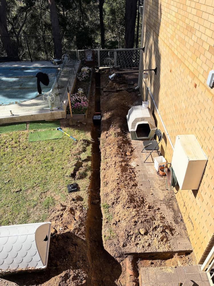 A Long Trench Dug Into the Ground Next to a Yellow Brick House — Crystal Clear Plumbing & Roofing in Wollongong, NSW