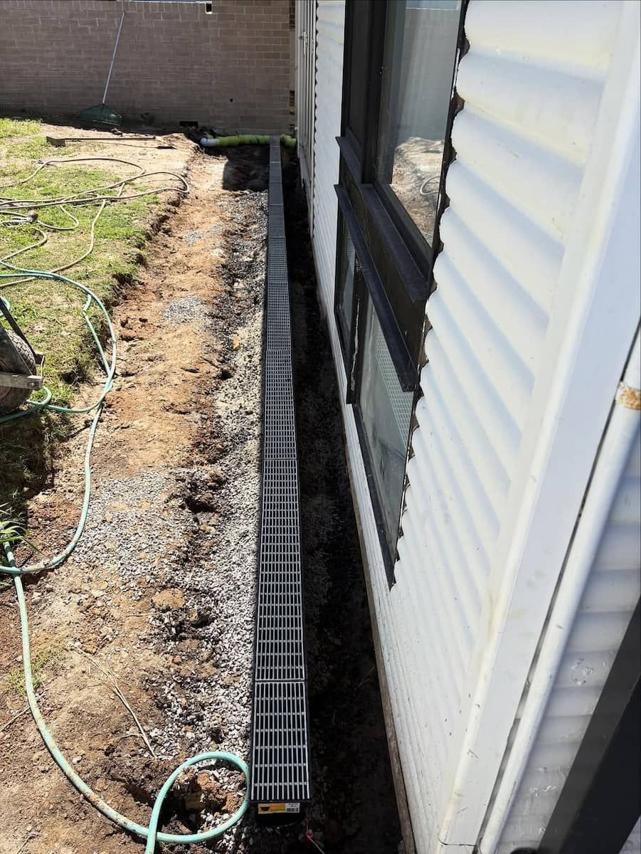 A Black Drainage Channel Installed in a Trench Beside a White House — Crystal Clear Plumbing & Roofing in Wollongong, NSW