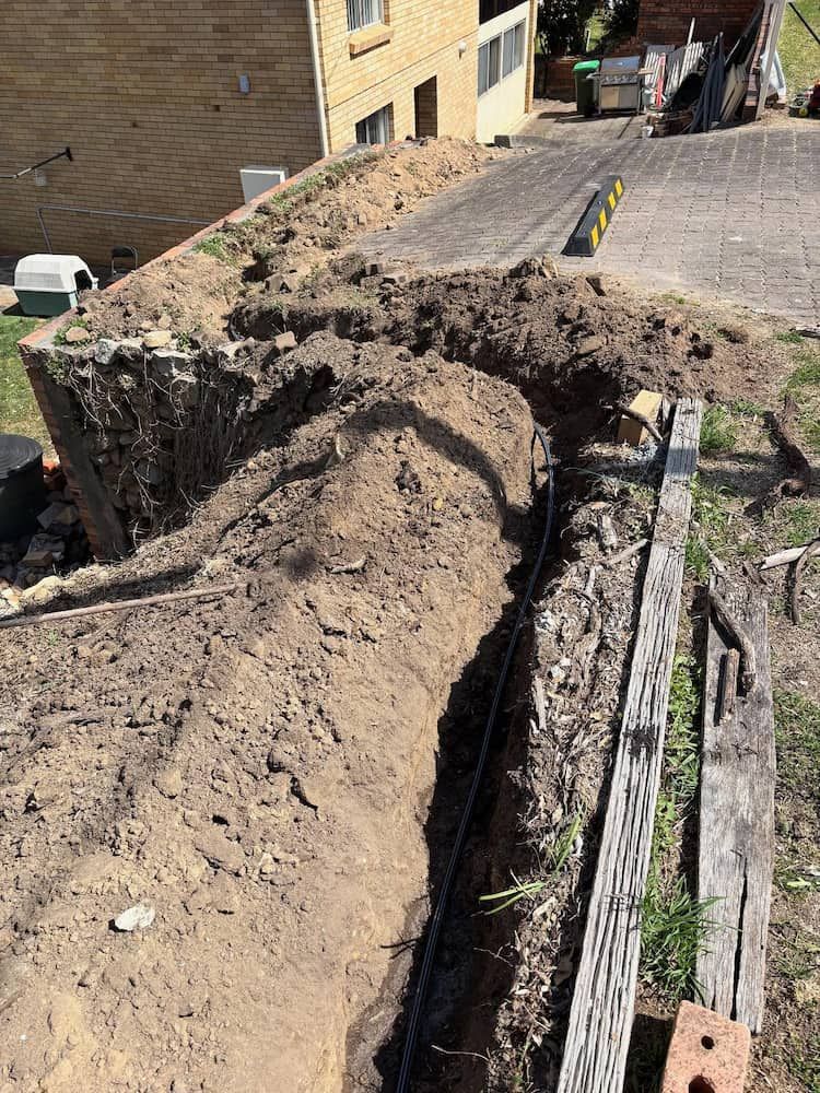A Trench Dug Into Soil Alongside a Wooden Sleeper Retaining Wall — Crystal Clear Plumbing & Roofing in Wollongong, NSW