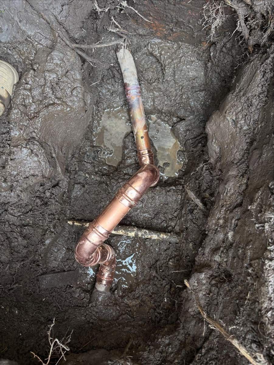 A repaired copper water pipe with several joints installed in a muddy, excavated dirt hole — Crystal Clear Plumbing & Roofing in Wollongong, NSW
