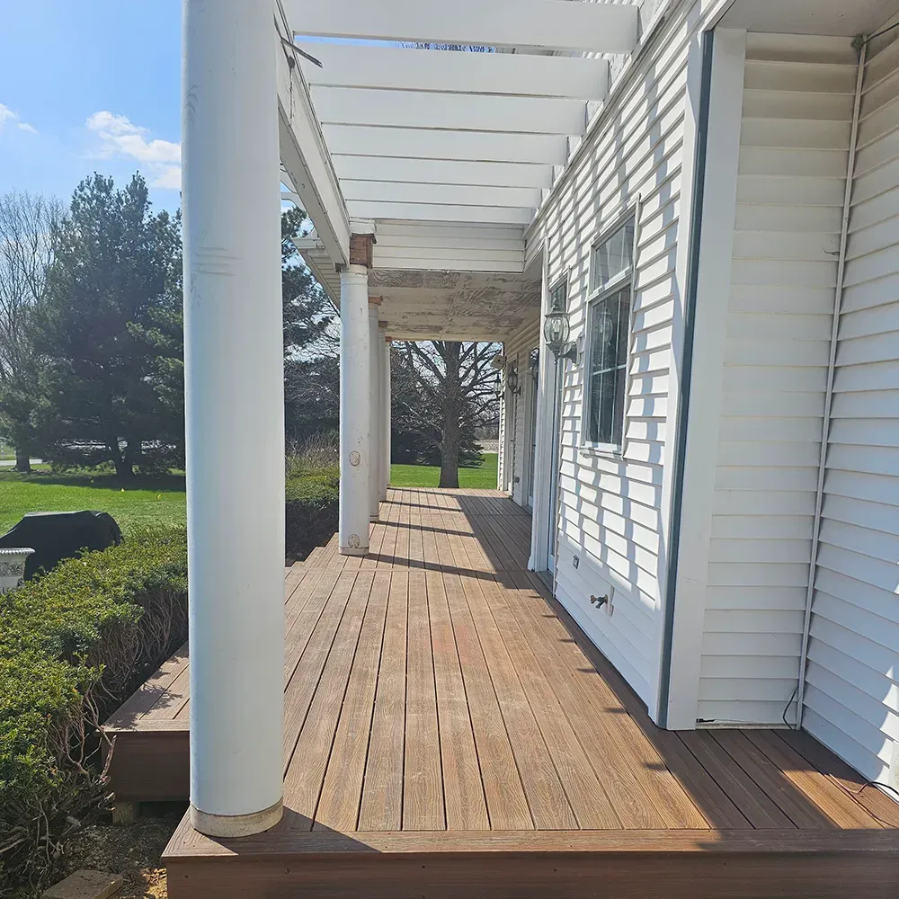 A wooden porch with white columns and a pergola roof extending along the side of a white-sided house on a sunny day.
