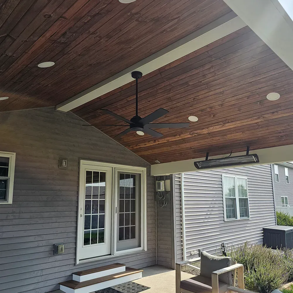 A covered patio with a wooden ceiling, ceiling fan, and mounted outdoor heater, attached to a house with gray siding.
