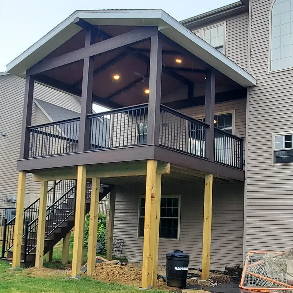 A raised wooden deck with a covered roof, black railings, and an outdoor staircase attached to a tan-sided house.