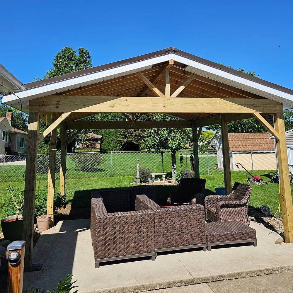 A wooden-framed outdoor pavilion with a dark roof covering wicker patio furniture on a concrete patio in a grassy yard.