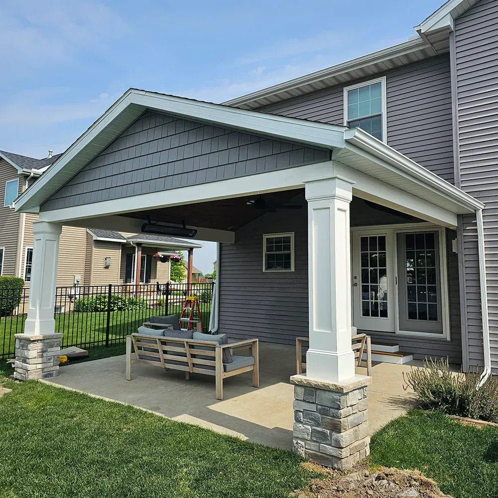 A gray house with a covered stone and white pillar patio featuring an outdoor wooden sofa, set in a backyard.