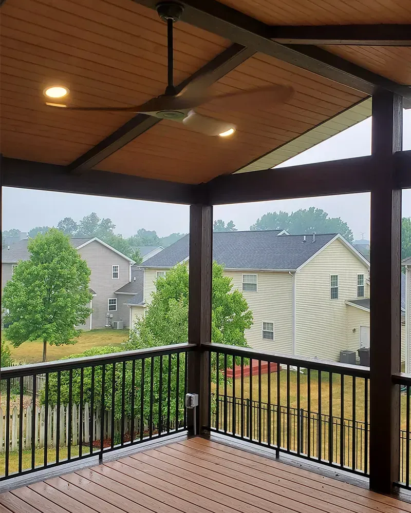 An elevated, covered wooden deck with a black railing, ceiling fan, and recessed lighting overlooking neighborhood homes.