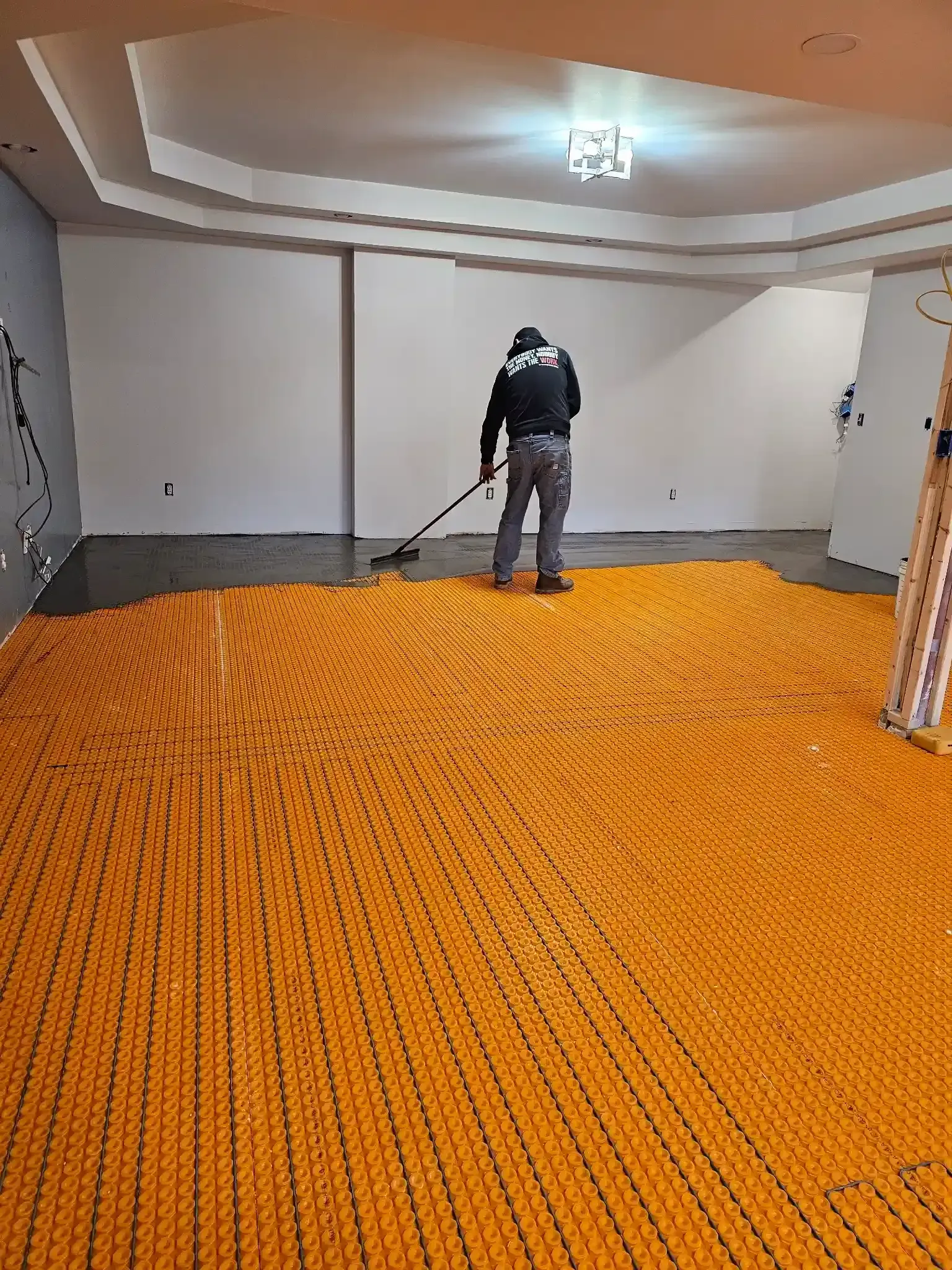 A worker uses a long-handled tool on a subfloor covered in an orange, grid-patterned underlayment membrane.