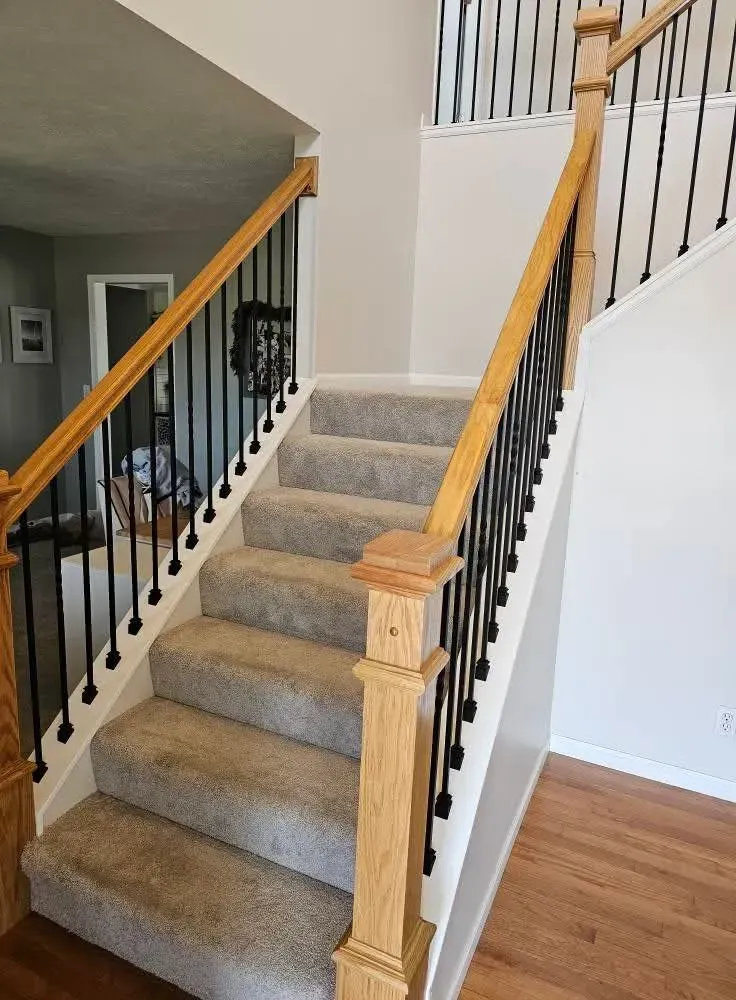 A staircase with gray carpet, light oak wood handrails and newel posts, and black metal balusters in a brightly lit hallway.