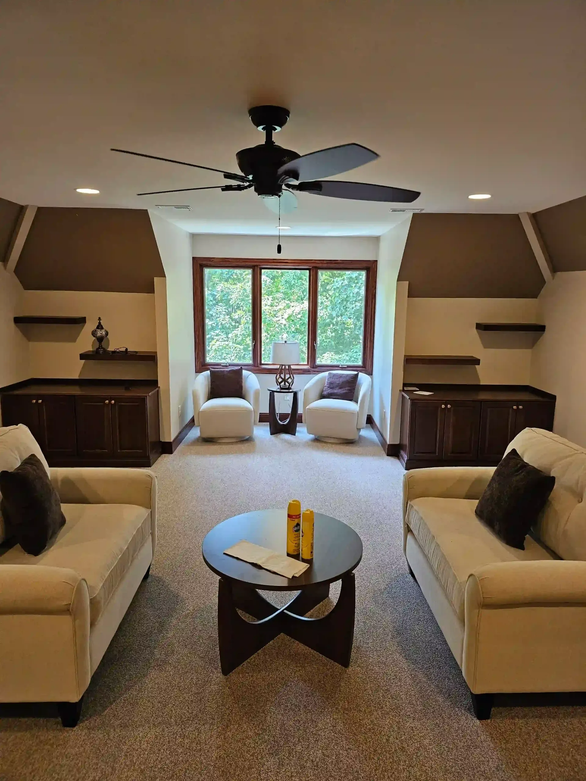 A carpeted living room featuring two beige sofas, two matching armchairs by a window, and a round dark coffee table.