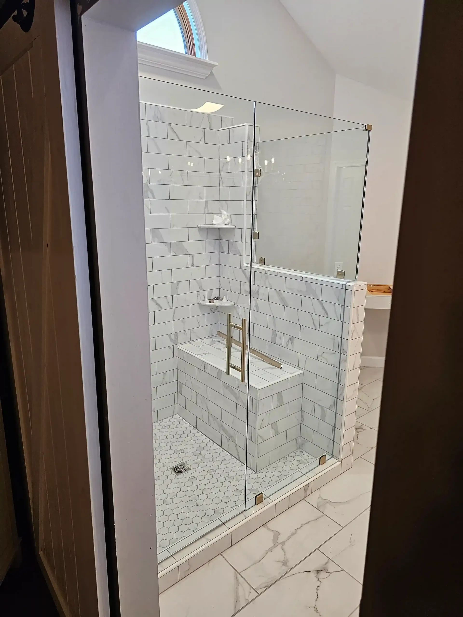 A walk-in shower with white and gray marble-patterned tiles, a built-in bench, glass doors, and gold-toned hardware.