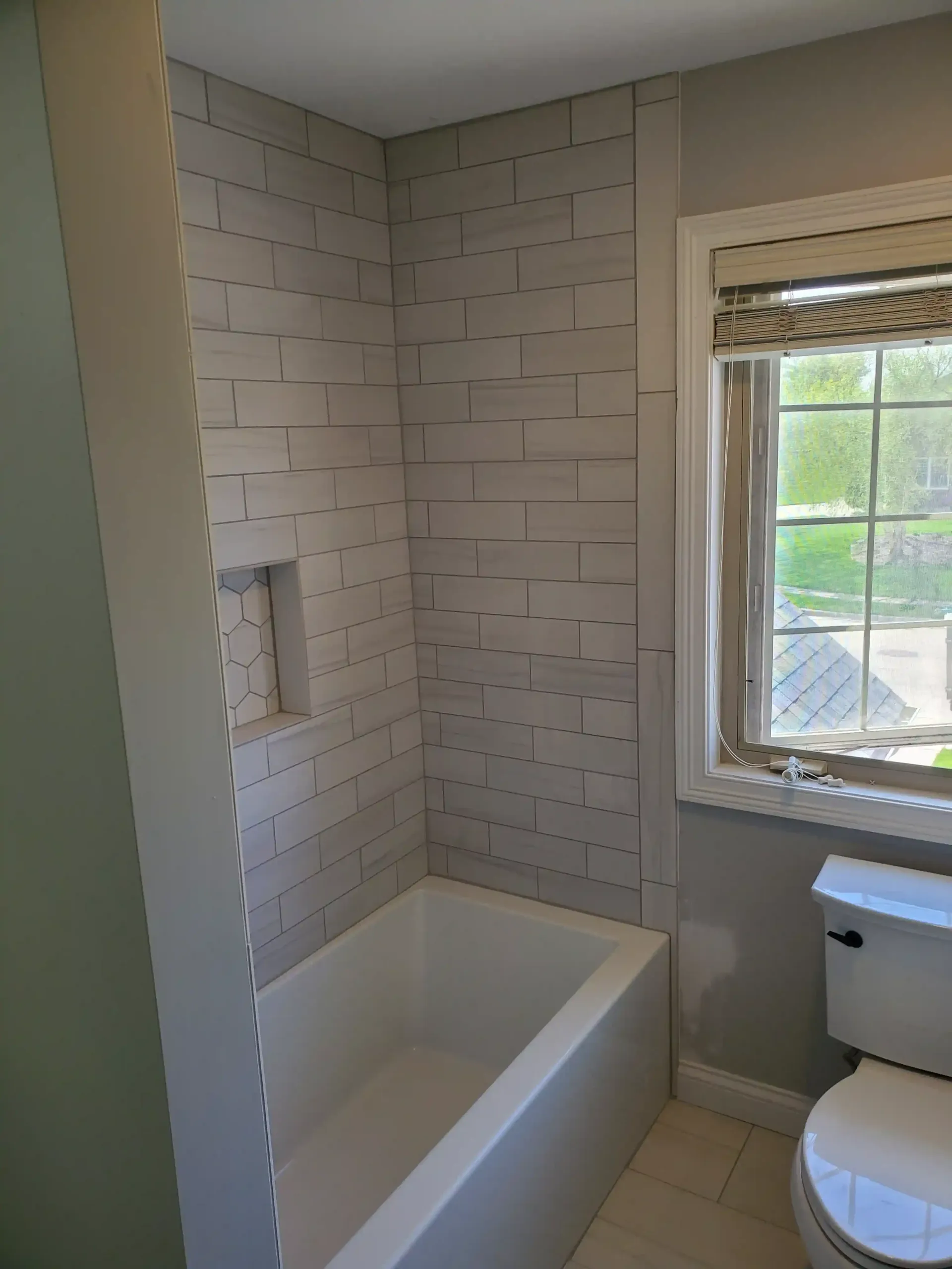A bathroom featuring a shower with light-colored brick-style tile, a white tub, and an adjacent toilet near a window.