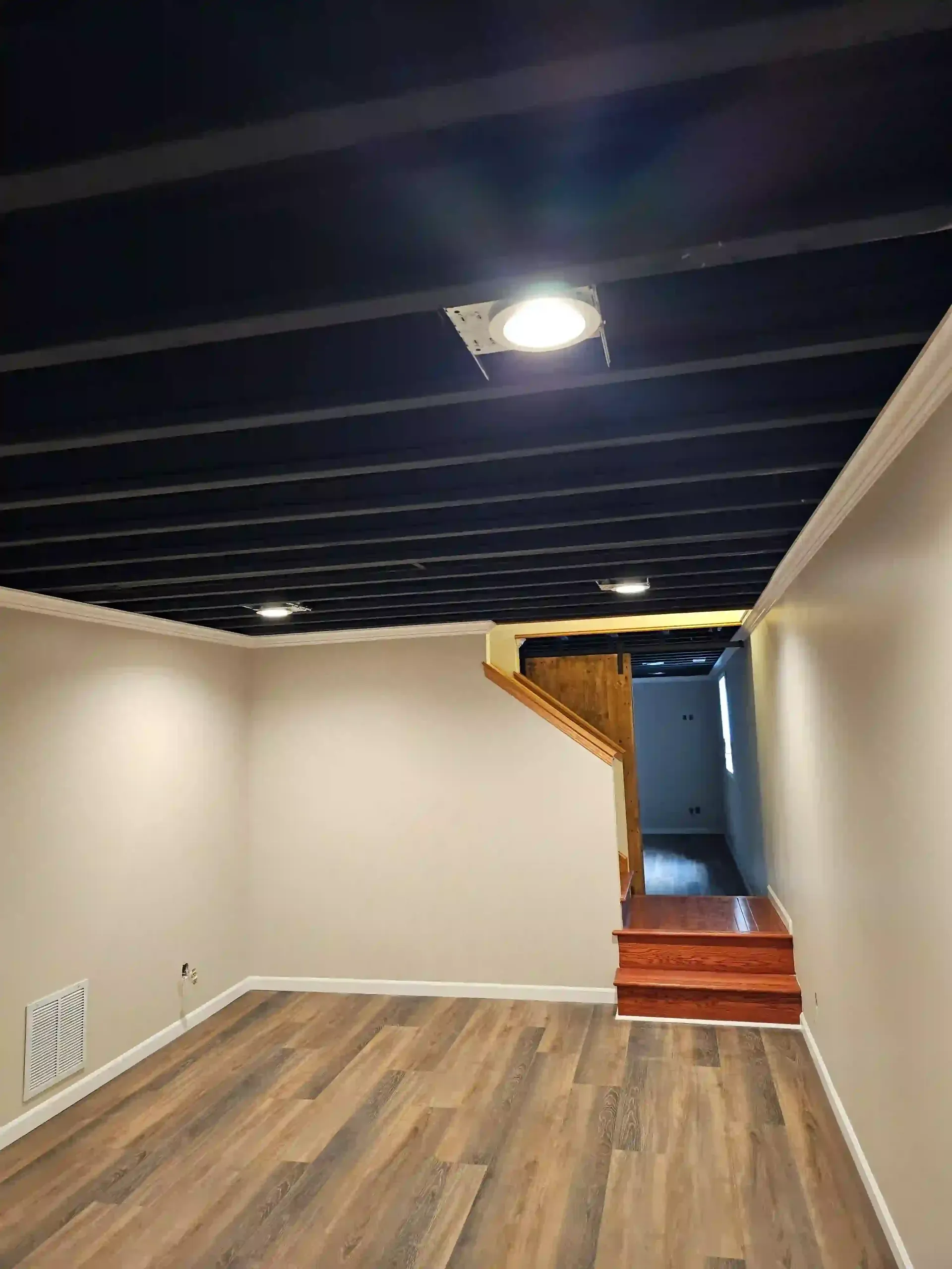 A basement with wood-look plank flooring, light beige walls, and an exposed ceiling painted black with recessed lighting.