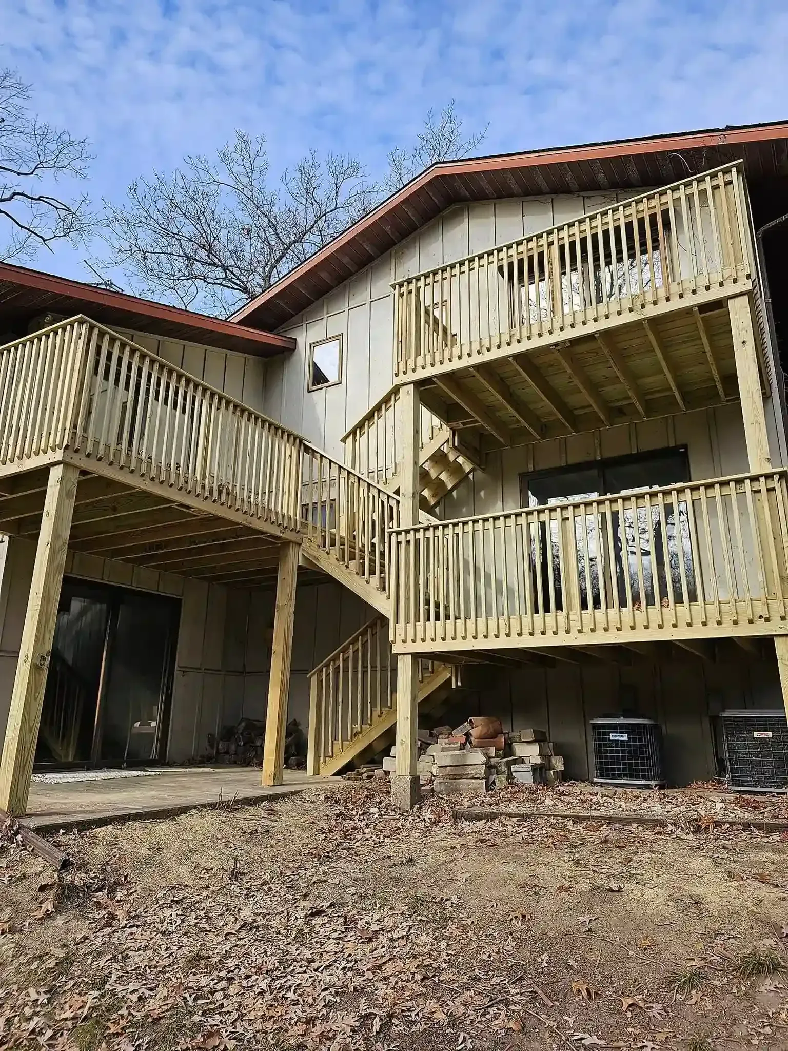 A two-story wooden deck with stairs connects to the back of a house over a yard covered in fallen leaves.