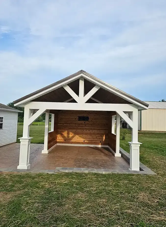 An open-air pavilion with a metal roof, white wooden support beams, and a natural wood interior wall on a paved patio.