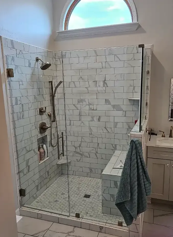 A modern walk-in shower with white marbled subway tile, glass walls, a built-in bench, and a rounded window above.
