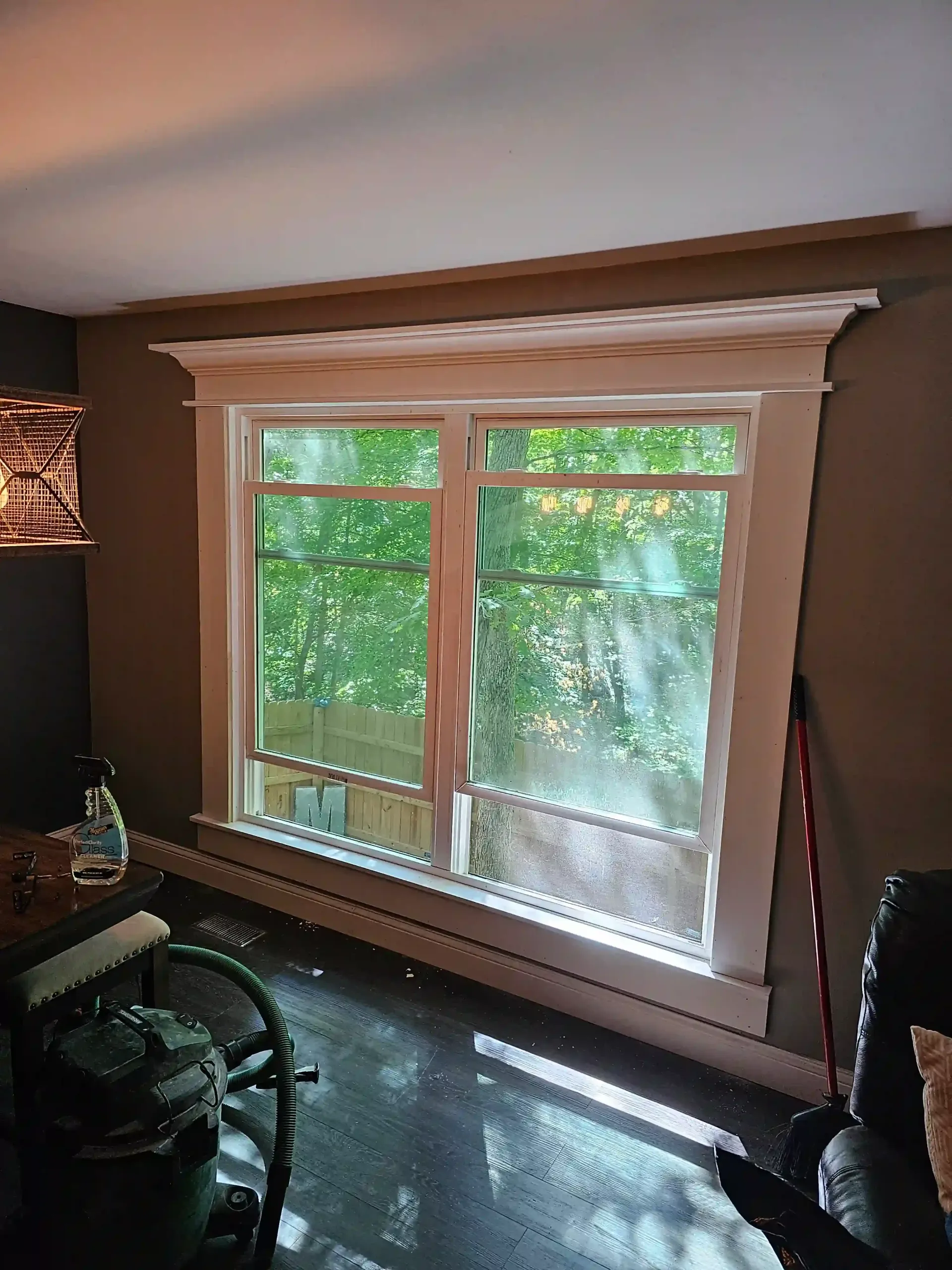 A large, white-trimmed window set into a dark brown wall, overlooking green trees outside, with a vacuum in the foreground.