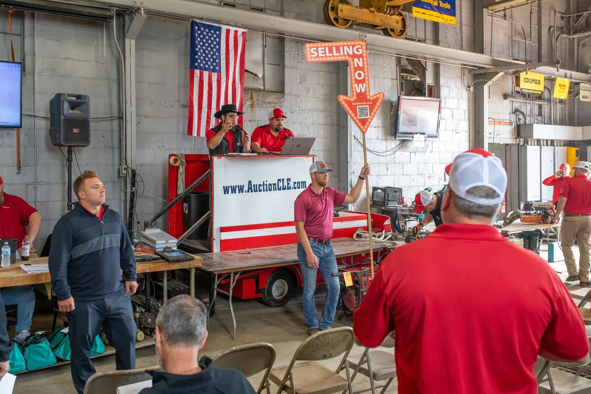 Auction in progress. People at an indoor equipment sale.  Sign pointing, selling now. American flag.  People wearing red.