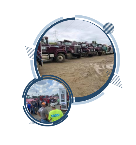 Line of maroon semi-trucks parked on dirt with people gathered at a truck show.