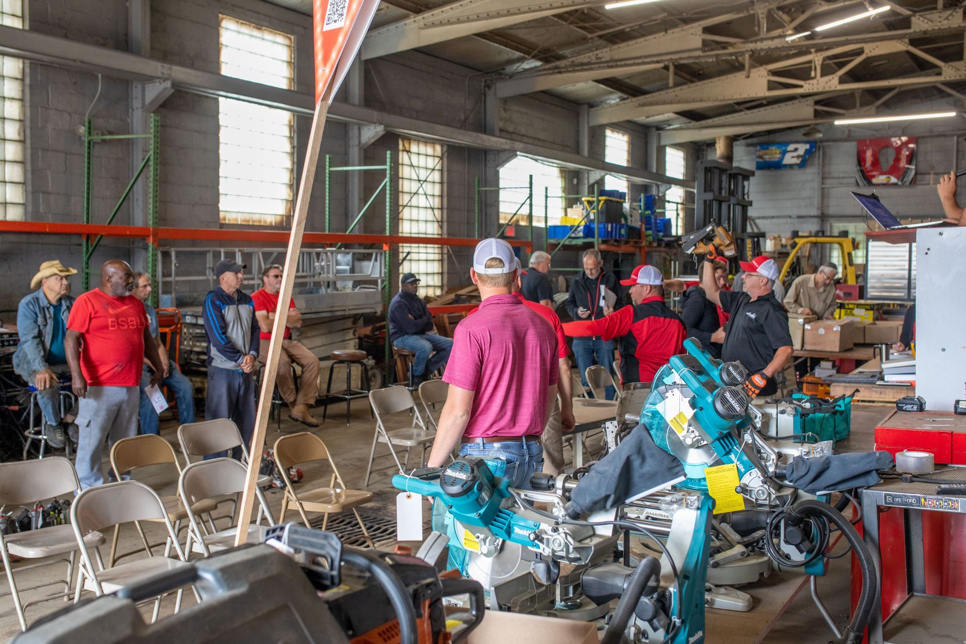 People at an auction in a warehouse; items on display, some seated, some standing, watching.