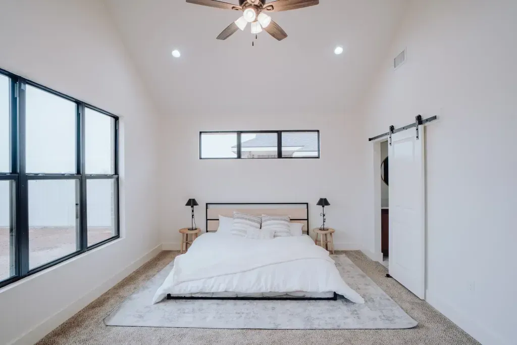 Bedroom with white walls, large windows, ceiling fan, bed, and sliding door.