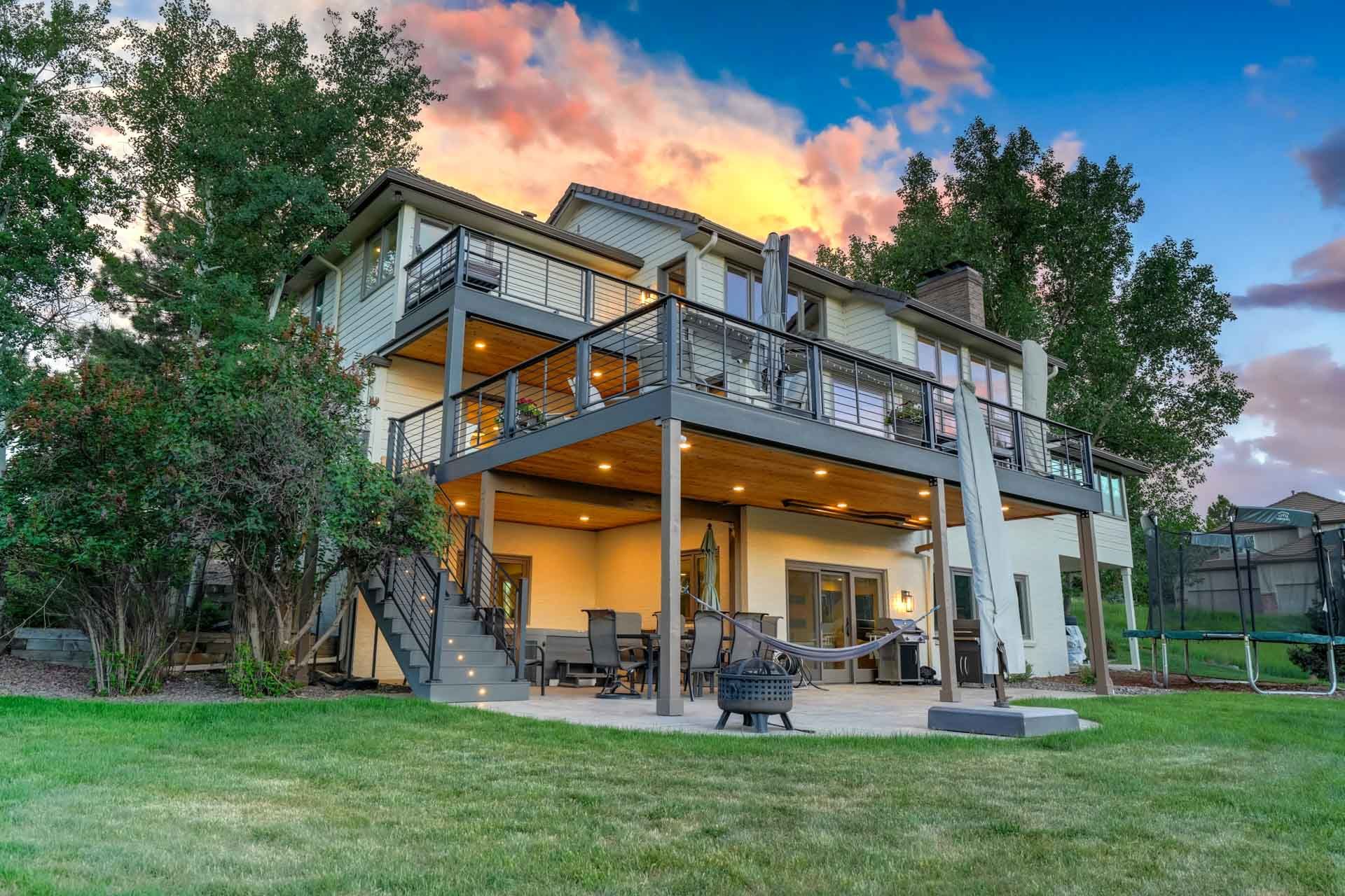 Modern two-story house with a deck and patio at sunset, surrounded by trees and a green lawn