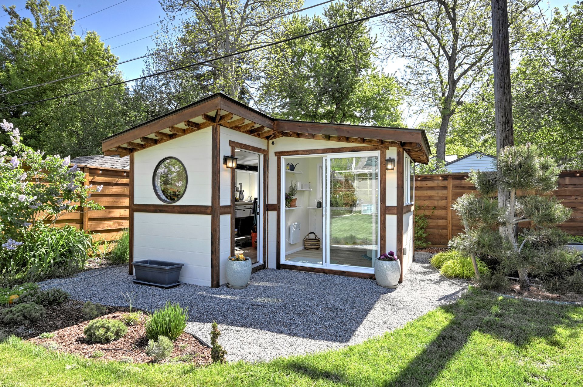 Modern backyard garden studio with glass sliding door, white walls, and gravel patio surrounded by trees.