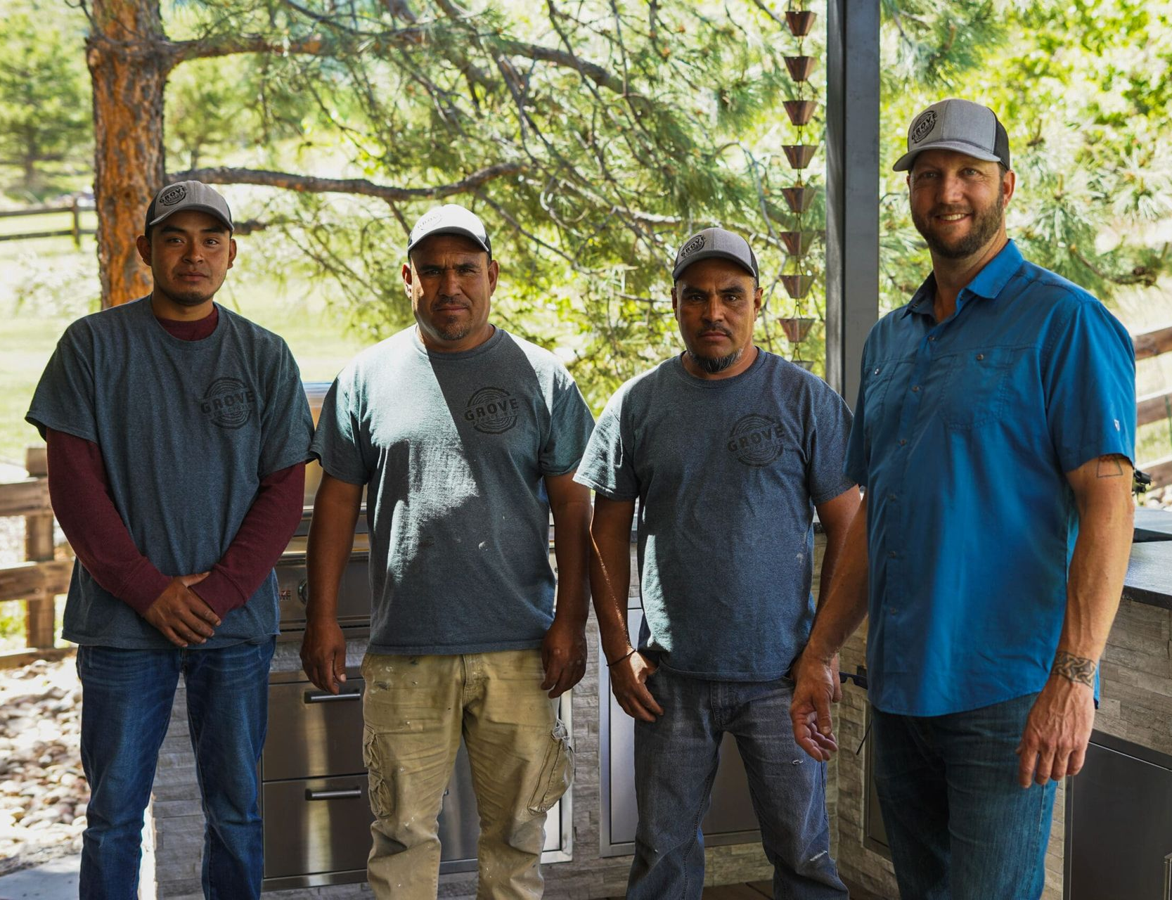 Four men stand side by side outdoors under trees, wearing casual shirts and caps, with a wooden structure behind them.