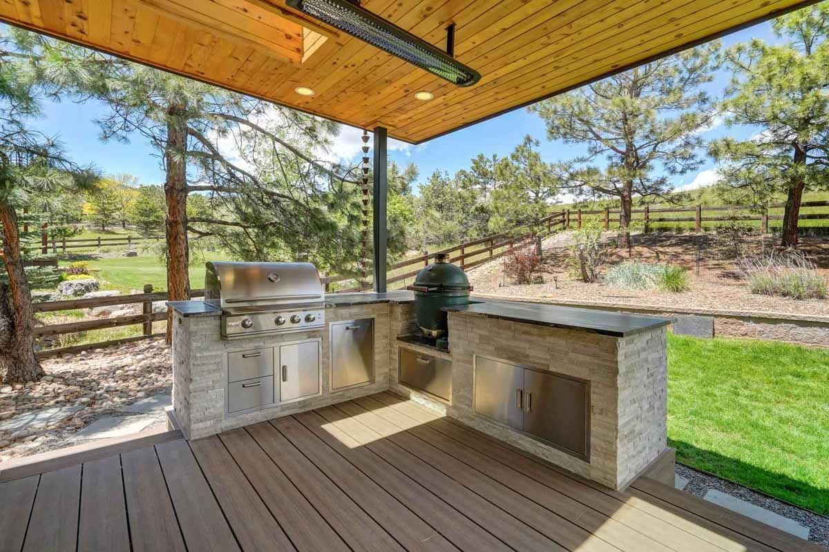 Covered outdoor kitchen with stainless grill, stone counters, and picnic area overlooking a grassy fenced yard.