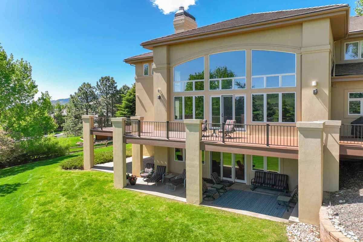 Large beige house with a wooden deck, tall windows, and a green lawn under a blue sky