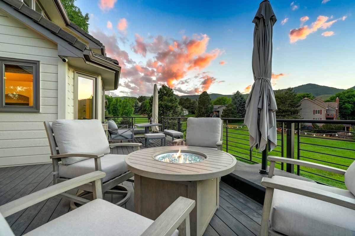 Patio with white chairs, round fire table, and closed umbrella overlooking a green backyard at sunset