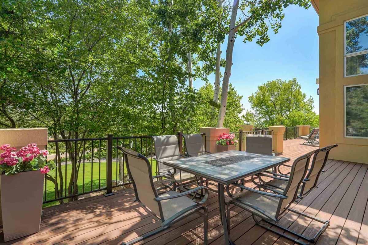 Patio with metal table and chairs beside flower planters, overlooking green trees on a sunny day