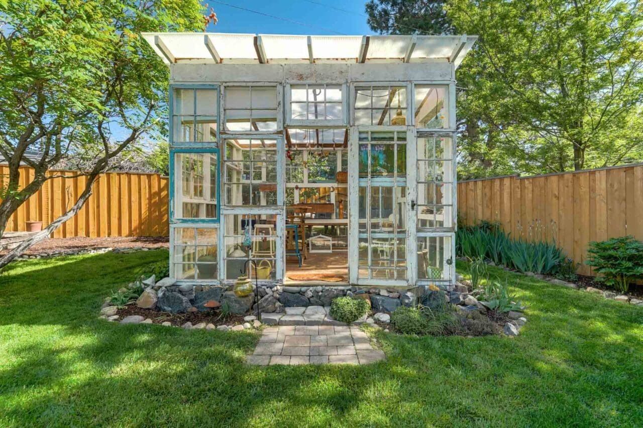 Glass greenhouse in a sunny backyard, with a stone path, green lawn, and wooden fence.