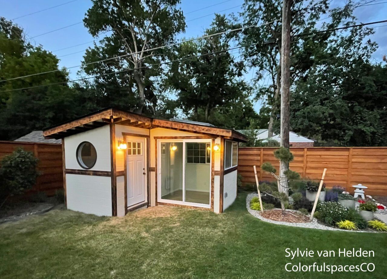 Small white garden shed with warm lights, wood trim, and glass doors in a backyard at dusk