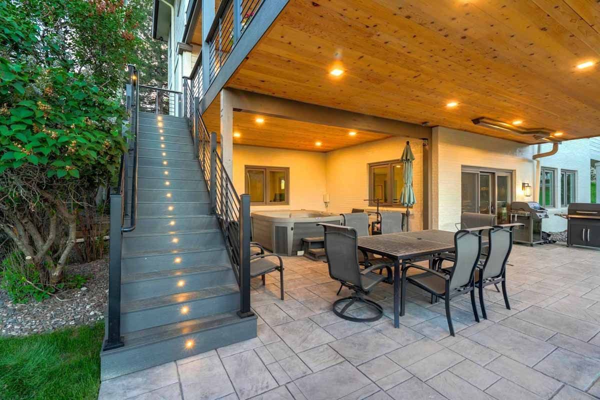 Covered patio with outdoor dining table, stairs, and warm lighting beside a house