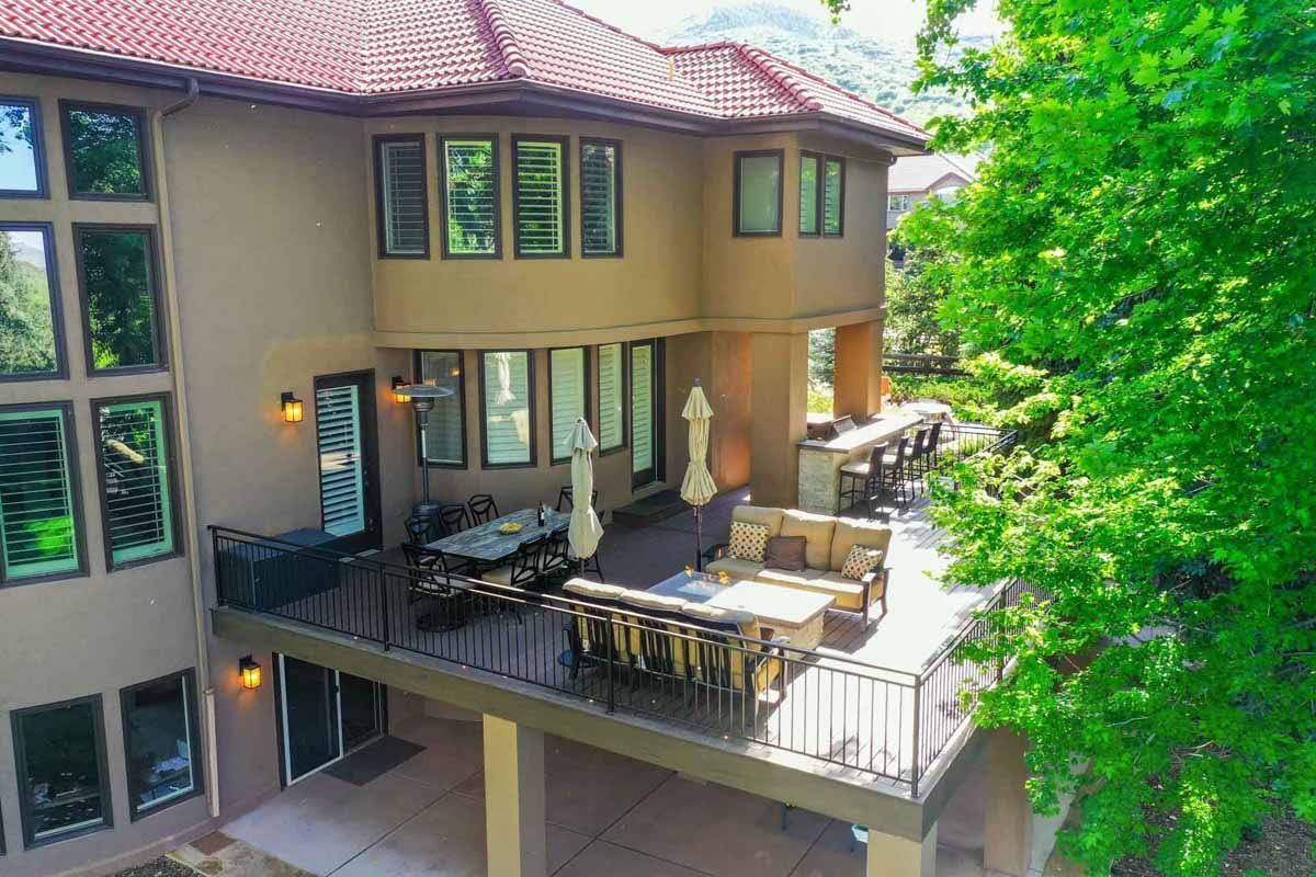 Two-story house with a patio deck, outdoor seating, and large windows surrounded by green trees