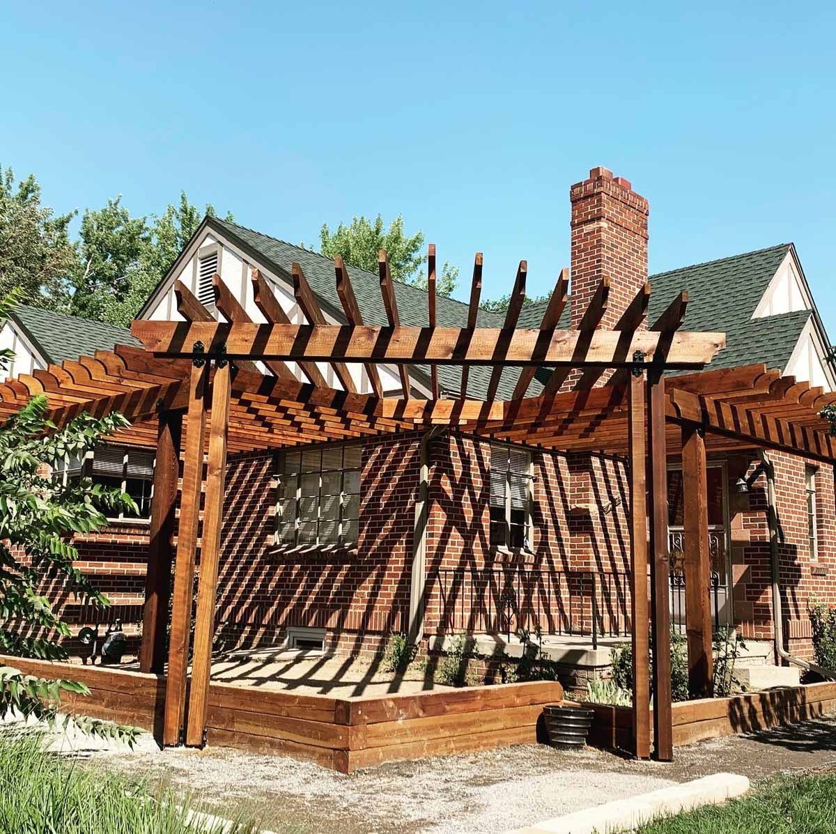 Wooden pergola on a brick patio beside a house with a red chimney and green roof under a blue sky