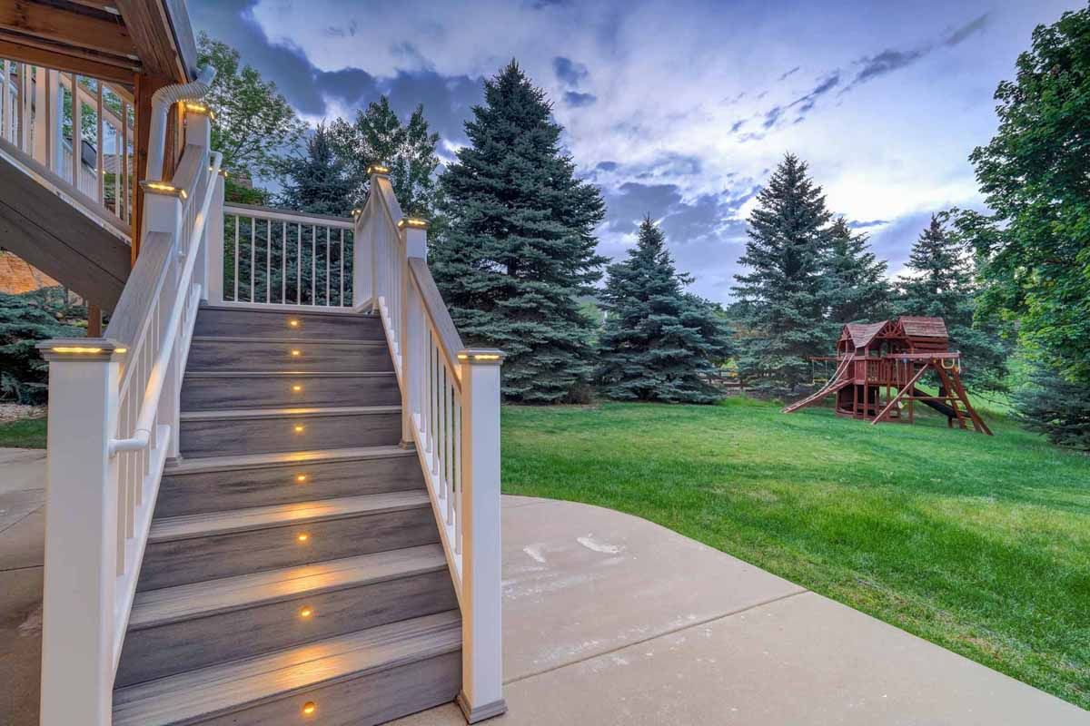 Lit stairs leading to a backyard lawn with evergreen trees and a red wooden gazebo at dusk