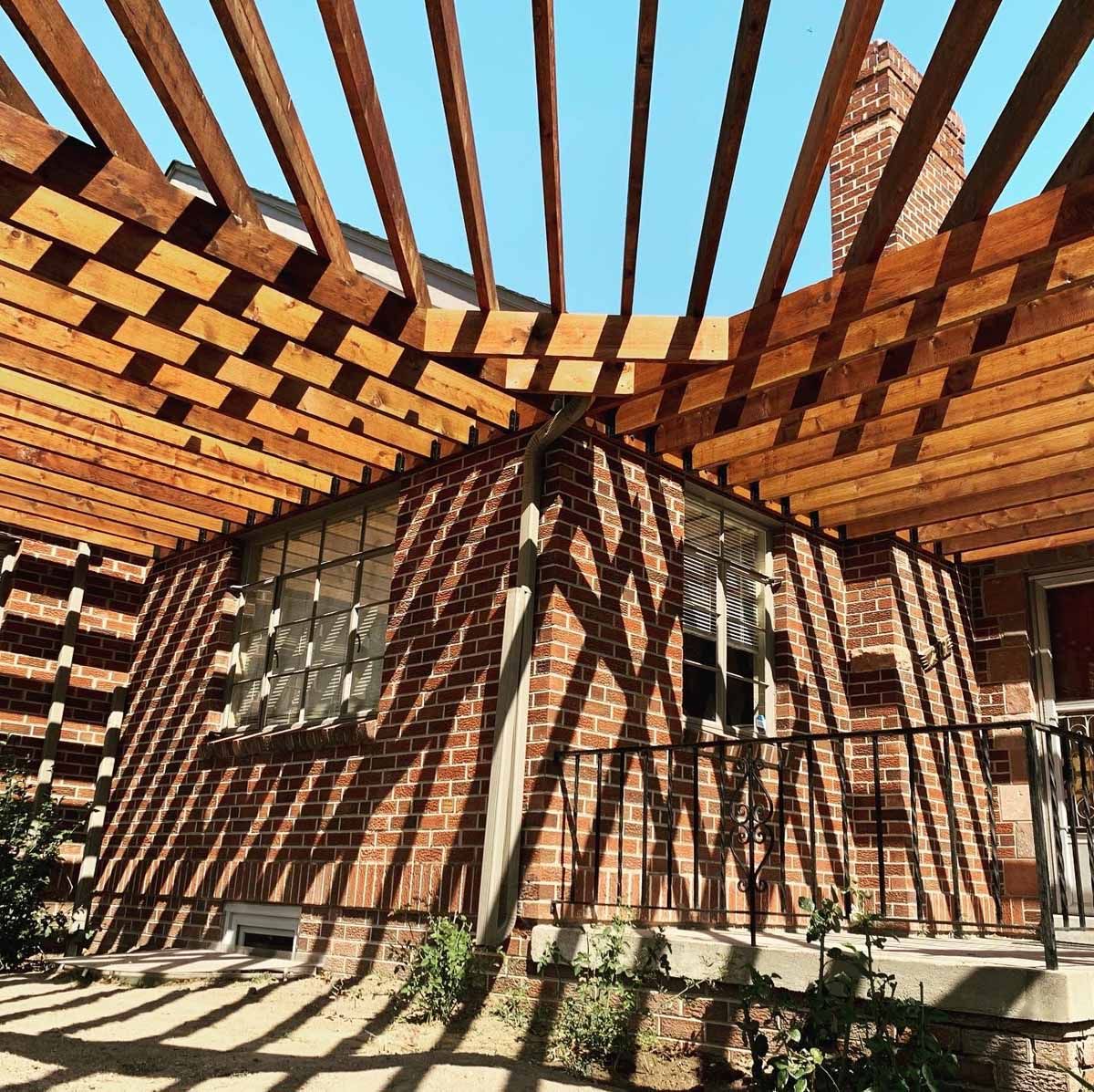 Brick house patio with wooden pergola beams casting striped shadows in bright sunlight